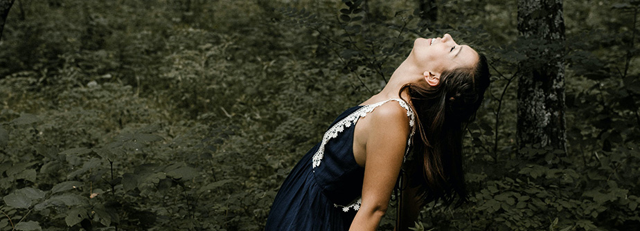 A young woman with a joyful expression tilts her head back in a dense green forest, wearing a dark dress with white lace trim.